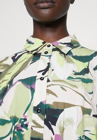 Ruusukuvioinen napitettava paita, jossa on vihreä, violetti ja kerma väriyhdistelmä, muotoiltu kaulus ja mustat napit teksturoidulla kankaalla.