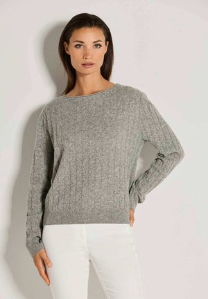 Frau mit dunklem Haar, die einen grauen Strickpullover und weiße Hose trägt, steht vor einer schlichten hellen Wand.