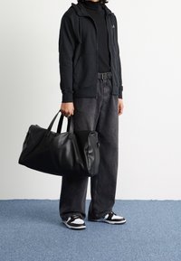 Svart läderduffelväska med två handtag, strukturerad form, slät textur och dragkedja. Bärs med mörk hoodie och baggy jeans.