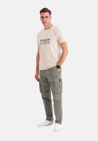 Beige bomulds T-shirt med sort tekst, parret med grønne cargo bukser med flere lommer. Modellen står i hvide sneakers.