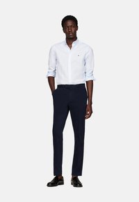 Camisa de botón azul claro con un pequeño logo, combinada con pantalones oscuros de corte slim y zapatos negros pulidos.