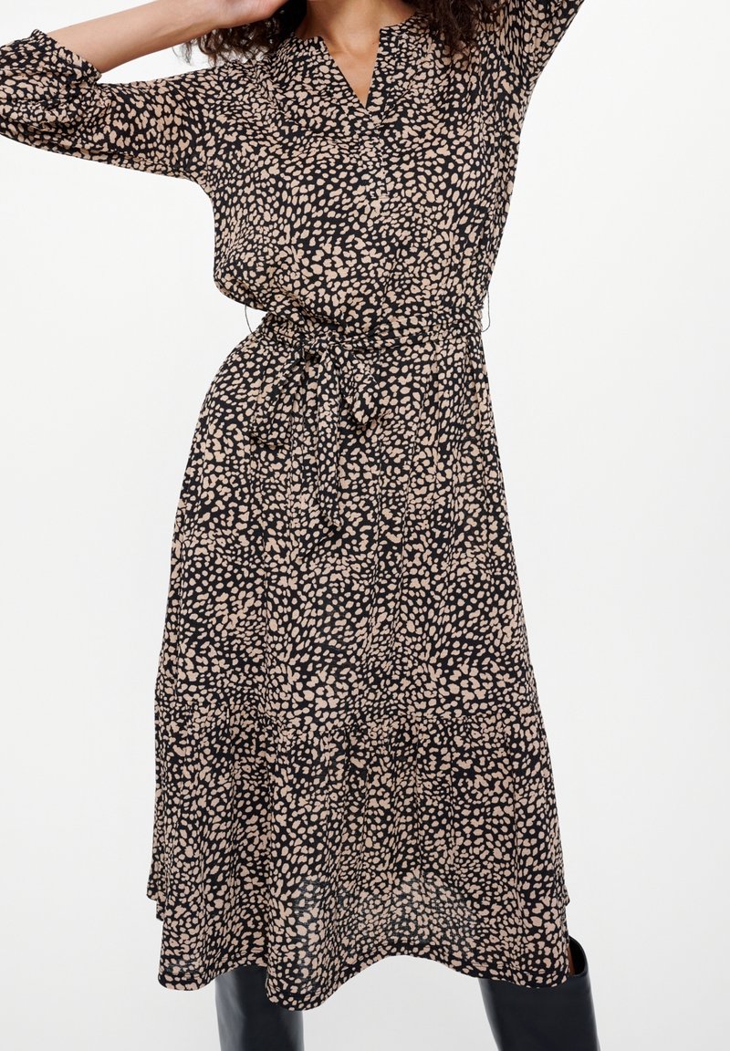 Žena v černých midi šatech s béžovým nepravidelným puntíkovým vzorem a zavázaným látkovým páskem, stojící s rukama zdviženýma proti bílému pozadí.