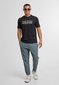 Černé tričko s krátkým rukávem a béžovým logem, spárované se světle šedými zúženými kalhotami a bílými teniskami, na jednoduchém pozadí.