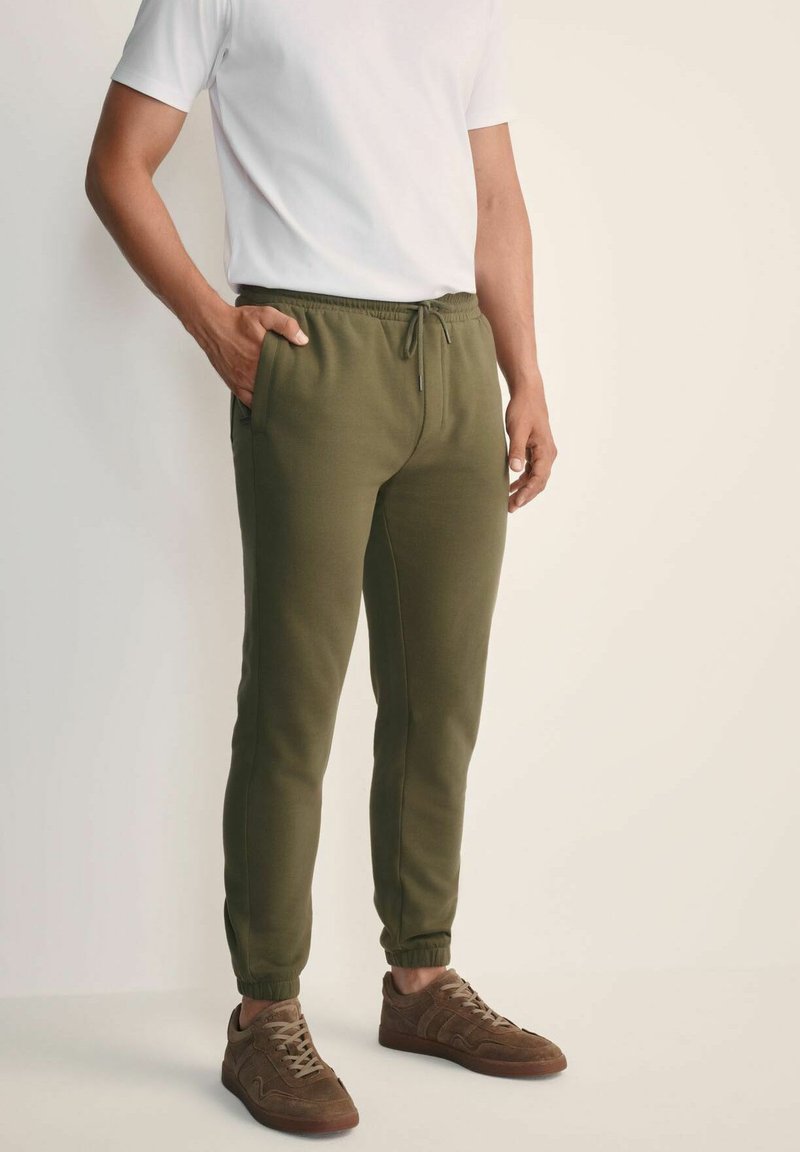 Mężczyzna w oliwkowych dresowych spodniach, brązowych sneakersach i białej koszuli z krótkim rękawem, stojący z jedną ręką w kieszeni na jednolitym tle.