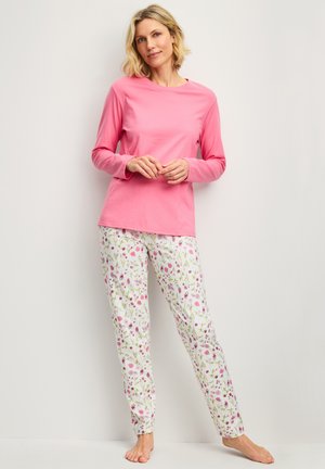 Woman standing barefoot wearing a long-sleeve pink top and white pajama pants with a multicolored floral pattern against a plain background.