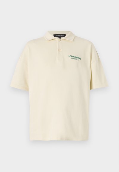 Polo en coton crème clair avec une coupe décontractée, manches courtes, trois boutons et texte brodé vert sur la poitrine.