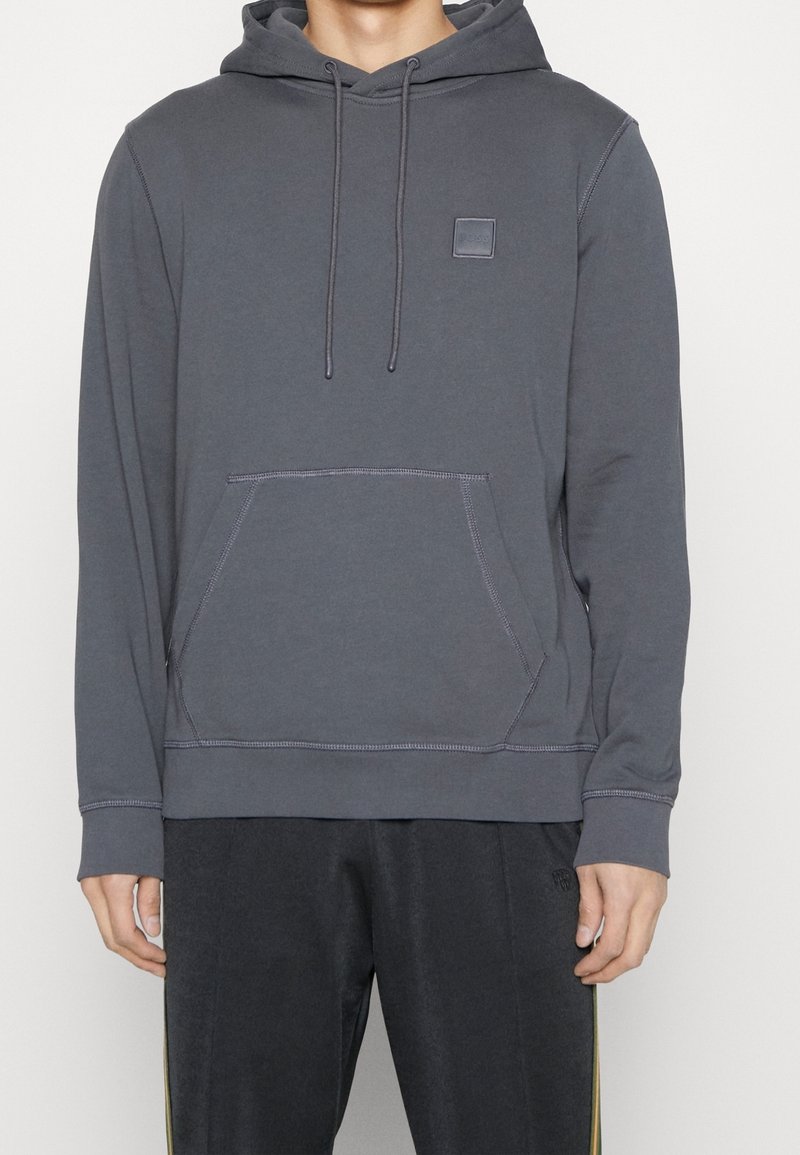 Sudadera gris de tela suave con bolsillo canguro, capucha con cordón y parche cuadrado en el pecho. Presenta detalles de costura en contraste.