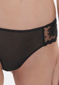 Czarne, przezroczyste majtki w stylu bikini z kwiatowym koronkowym detalem na biodrach noszone na jasnej karnacji.