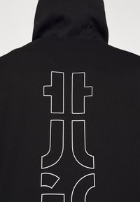 Sort hættetrøje med et stort hvidt grafikdesign på ryggen, bestående af fede abstrakte former og bogstaver. Glat stofstruktur.