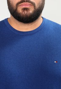 Homme barbu portant un pull bleu à col rond avec un petit logo rectangulaire rouge, blanc et noir sur la poitrine.
