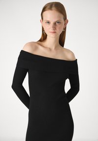 Svart, ribbestrikket avskjært kjole med lange ermer. Stoffet har en teksturert overflate med vertikale linjer som fremhever en tettsittende silhuett.