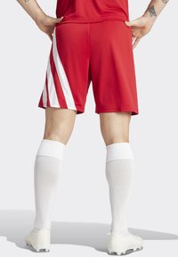 Rote Sportshorts mit weißen Seitenstreifen, kombiniert mit weißen Kniestrümpfen und hellen Fußballschuhen. Ansicht von hinten.