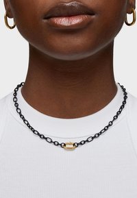 Collana in catena nera con anelli oblunghi e dettaglio della chiusura in oro. Indossata sopra una maglietta bianca a righe, esaltando i colori contrastanti.