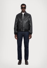 Chaqueta bomber de cuero negra con cierre de cremallera, puños y dobladillo acanalados. La lleva puesta sobre una camisa blanca y la combina con jeans azul oscuro y zapatos marrones.