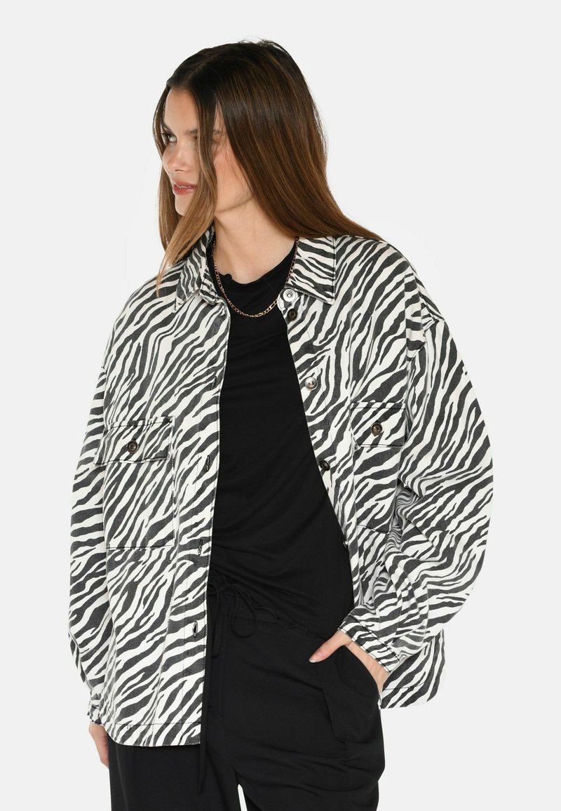 Zebratäpläkkjest i svart og kvitt, oversized design, laget av teksturert stoff, med to frontlommer og knappelukking.