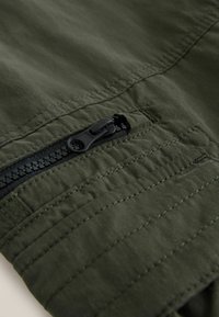 Close-up of dark green fabric with black zipper and detailed stitching on a pocket area.