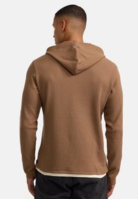 Pullover marrone con cappuccio realizzato in un tessuto testurizzato e a coste. Dotato di tasca a marsupio e orlo curvo. Design casual con maniche lunghe e cappuccio.