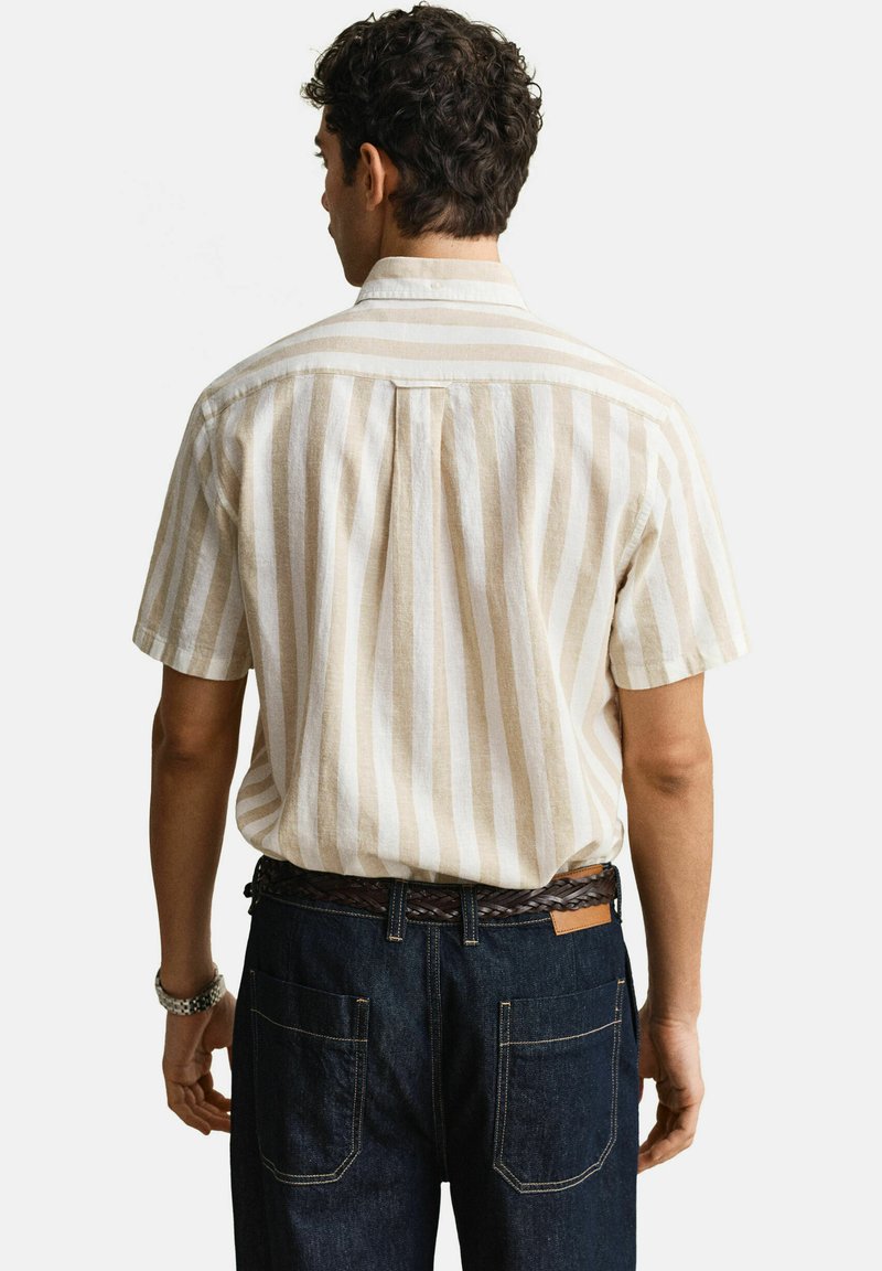 Mann, der ein beige-weiß gestreiftes Kurzarmhemd mit vertikalen Streifen trägt, in die dunkelblauen Jeans mit einem braunen geflochtenen Gürtel gesteckt, von hinten gesehen.