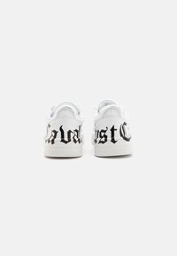 White sneakers featuring a smooth leather texture with black gothic script on the heel. Rounded toe and low-top design with lace-up closure.