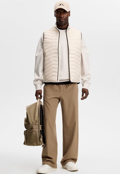 Uomo con cappellino beige, maglione bianco sporco, gilet trapuntato beige, pantaloni marroni, sneaker bianche, che tiene uno zaino beige di lato.