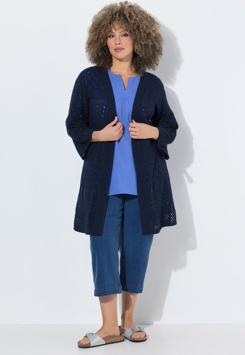 Woman with curly hair wearing a navy knit cardigan, blue top, cropped jeans, and silver sandals, standing against a white background.
