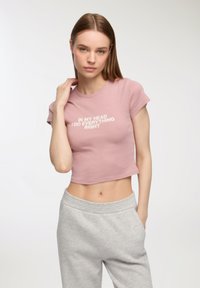T-shirt corta rosa chiaro con maniche corte e testo bianco che recita: "Nella mia testa faccio tutto giusto." Abbinata a pantaloni grigi.