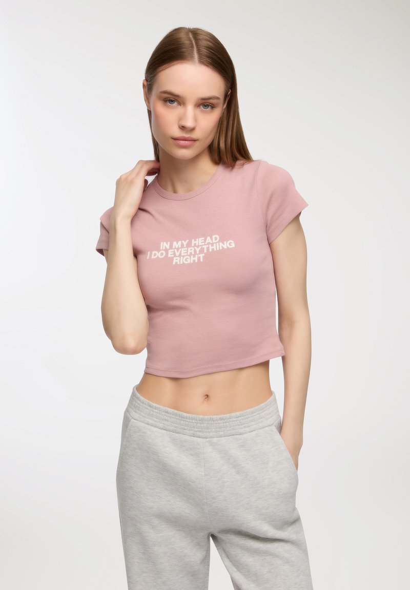 T-shirt corta rosa chiaro con maniche corte e testo bianco che recita: "Nella mia testa faccio tutto giusto." Abbinata a pantaloni grigi.