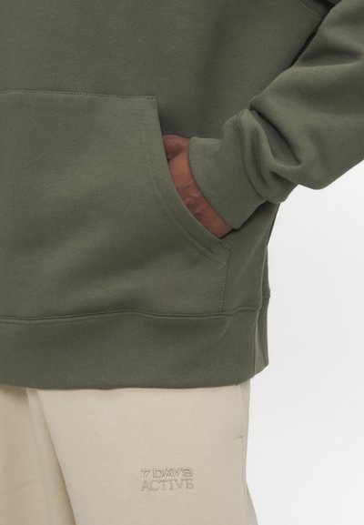 Hanorac verde măsliniu cu buzunar frontal și manșete cu nervuri, asortat cu pantaloni sport bej cu branding brodat.