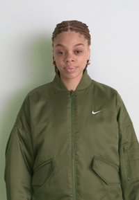 Maslinasto zelena bomber jakna sa zatvaračem sprijeda, bočnim džepovima i Nike swoosh logom. Napravljena od glatkog, laganog materijala.