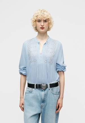 Plavovlasá osoba s kučeravými vlasmi, ktorá nosí svetlomodrú vyšívanú blúzku s vyhrnutými rukávmi a svetlomodré rifle s vysokým pásom s opaskom so srdcovou prackou.