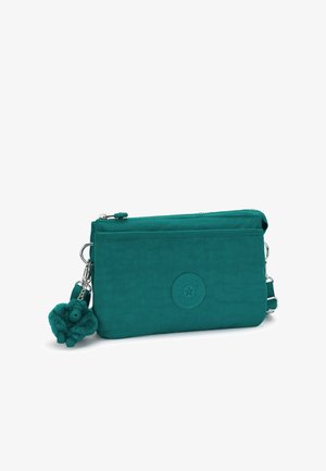 Obdélníková tyrkysová crossbody taška s předním zip kapsou, nášivkou s logem Kipling a připevněným plyšovým klíčenkovým opičkou na bílém pozadí.