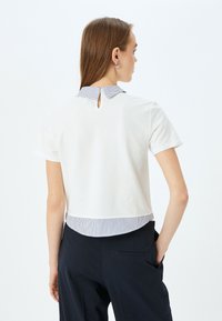 T-shirt bianco a maniche corte con colletto a righe, dotato di un dettaglio a chiave sulla schiena e orlo a righe sul fondo. Il materiale sembra morbido.