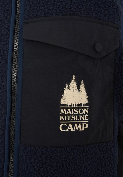 Marineblå fleece-jakke med teksturert overflate, med en flat lomme og kremfarget logo av trær og teksten "MAISON KITSUNÉ CAMP." Glidelås foran.