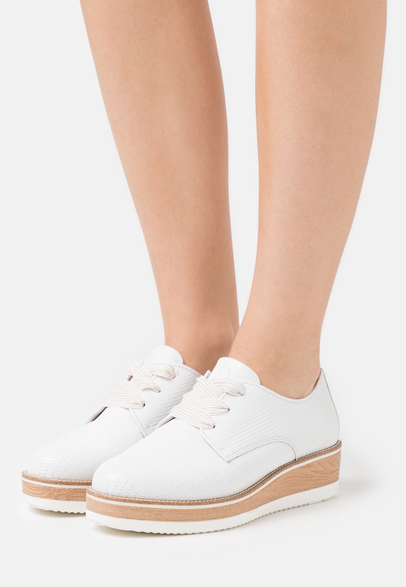 Chaussures à plateforme en cuir blanc à lacets avec semelles épaisses beige clair, portées pieds nus, présentées sur un fond blanc uni.