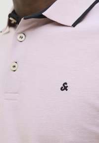 Polo skjorte i lys pink med struktureret stof, sorte krave accenter og tre knapper. Har et lille sort broderet logo på brystet.