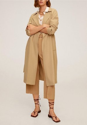 Kvinde iført beige trenchcoat, matchende cropped bukser, hvid bluse, lagdelte halskæder og brune snøresandaler, stående med armene krydsede.