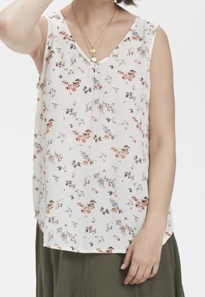 Kvinde iført hvid ærmeløs bluse med lille blomsterprint, lagdelte gyldne halskæder og olivengrøn nederdel mod ensfarvet baggrund.