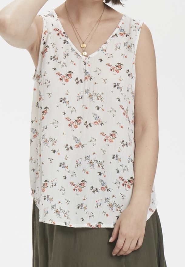 Femme portant un chemisier blanc sans manches à petit motif floral, des colliers dorés superposés et une jupe vert olive sur fond uni.