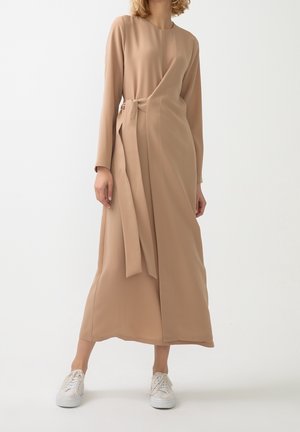 Eine Frau trägt ein langes beiges Wickelkleid mit seitlicher Schleife und weißen Turnschuhen und steht vor einem schlichten weißen Hintergrund.