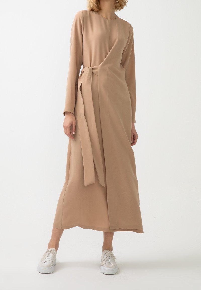 Femme portant une longue robe portefeuille beige avec un nœud sur le côté et des baskets blanches, debout devant un fond blanc uni.