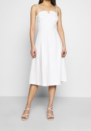 Femme portant une robe midi blanche sans manches avec de fines bretelles et des sandales blanches à talons croisées sur un fond uni.