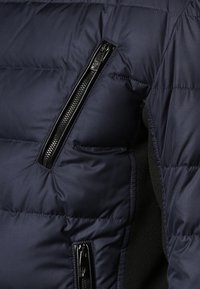 Mörkblå puffjacka med horisontell vaddering, svarta dragkedjor och en sidficka. Materialet är slätt med en lätt glänsande yta.