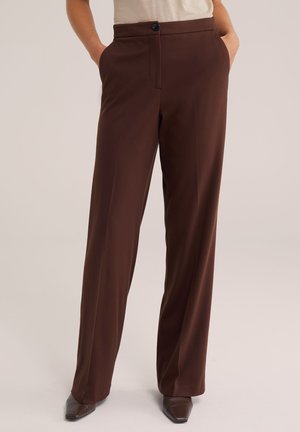 Persona que lleva pantalones marrones de talle alto y pierna ancha con bolsillos laterales y zapatos marrones de punta afilada sobre un fondo liso.