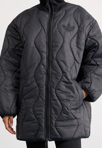 Chaqueta de plumas acolchada negra con un alto cuello, que cuenta con una cremallera frontal completa, patrones de ondas texturizadas y un pequeño logo bordado en el pecho.