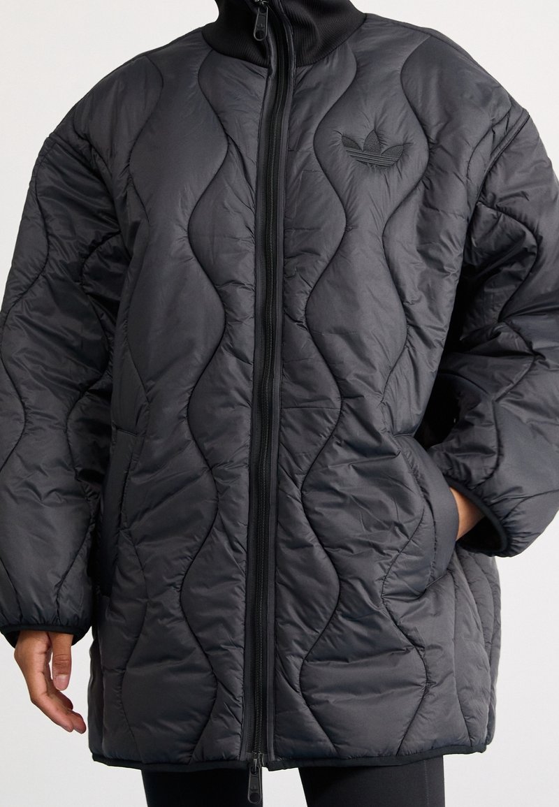 Chaqueta de plumas acolchada negra con un alto cuello, que cuenta con una cremallera frontal completa, patrones de ondas texturizadas y un pequeño logo bordado en el pecho.