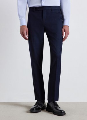 Mann trägt marineblaue Anzughose, hellblaues Hemd mit Knöpfen, schwarze Anzugschuhe, steht vor einem schlichten hellen Hintergrund.