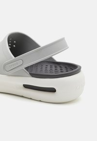 Sive gumene sandale s bijelim potplatom, s širokim remenom i ventilacijskim otvorima na ulošku. "Crocs" logo na remenu.