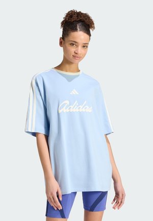 Jeune femme aux cheveux bouclés portant un t-shirt Adidas oversize bleu clair et un short de cyclisme bleu, debout devant un fond uni.