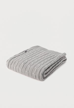 Hellgrauer, gefalteter Schal aus Kabelstrickwolle mit einem kleinen schwarzen Sticklogo an einer Ecke, auf weißem Hintergrund präsentiert.