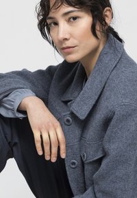 Abrigo de mezcla de lana gris con cuello, dos botones y solapas en los bolsillos. Combinado con una manga gris claro visible por debajo. Diseño simple y estructurado.